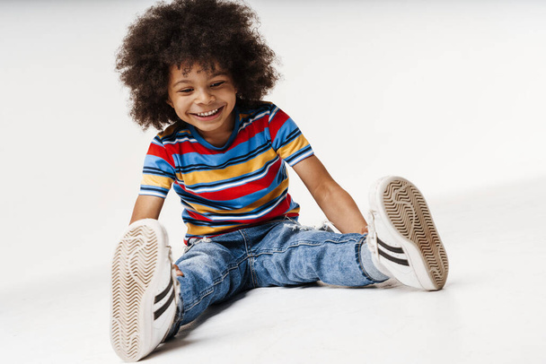Smiling little african boy in everyday clothes over wall background, sitting on a floor - Foto, Bild