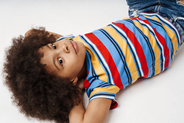 Little african boy in everyday clothes over wall background, laying on a floor - Фото, изображение