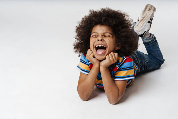 Smiling little african boy in everyday clothes over wall background, laying on a floor - Foto, immagini