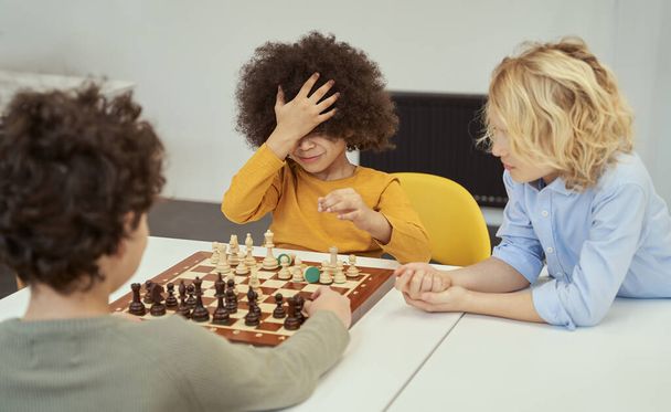 Oops. Funny little boy with afro hair made wrong move. He is touching his head as a sign of failure while sitting at the table and playing chess with friends indoors - Φωτογραφία, εικόνα