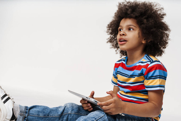 Preschooler african boy wearing t-shirt sitting over white wall, holding digital tablet computer, looking aside - Foto, imagen