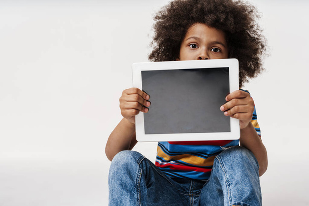 Preschooler african boy wearing t-shirt sitting over white wall, showing empty digital tablet computer - Zdjęcie, obraz