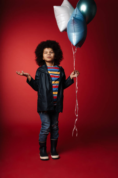 Full length of an african little kid holding bunch of balloons over red wall background - Φωτογραφία, εικόνα