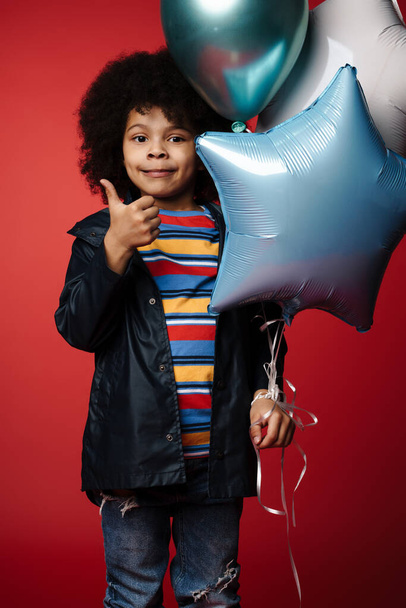 African little kid holding bunch of balloons over red wall background, thumbs up - Valokuva, kuva