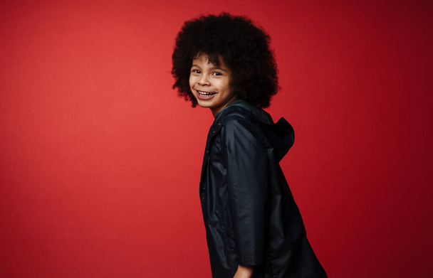 Happy preschooler african boy wearing coat standing over red wall, having fun, side view, looking camera - Photo, image