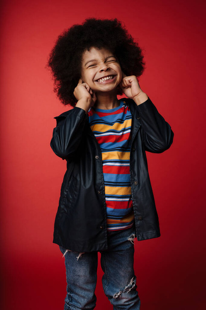 Happy preschooler african boy wearing coat standing over red wall, having fun, covers ears - 写真・画像