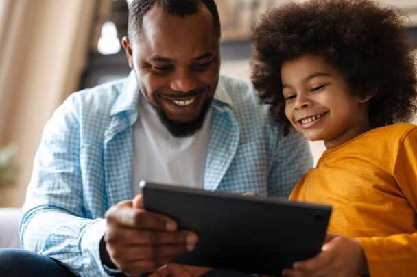 Black father and son smiling and using tablet computer while sitting on sofa at home - Photo, image