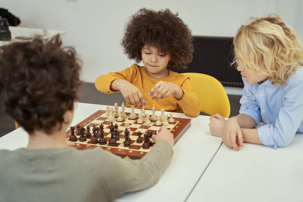 Impresionantes niños pequeños discutiendo juego mientras están sentados en la mesa y jugando al ajedrez en el interior - Foto, Imagen