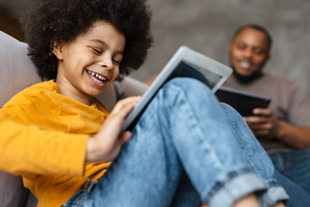 Black father and son smiling and using tablet computers while sitting on sofa at home - Foto, Imagem