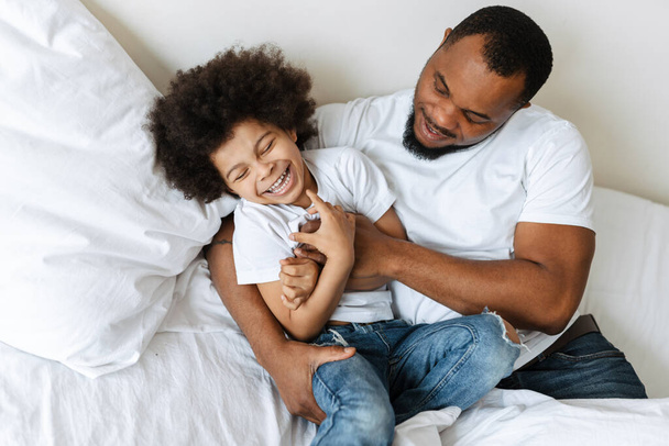 Black father and son laughing while making fun in bed at home - Valokuva, kuva
