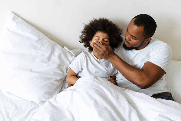 Schwarzer Vater bedeckt den Mund seines Sohnes, während er zu Hause im Bett liegt - Foto, Bild