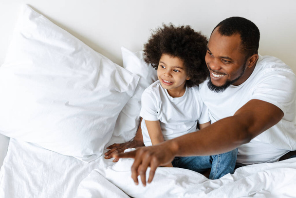 Black father and son smiling and looking aside while sitting in bed at home - Fotografie, Obrázek