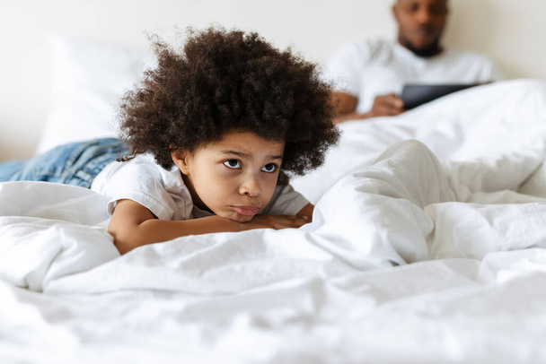 Black boy frowning while his father using tablet computer in bed at home - Fotó, kép