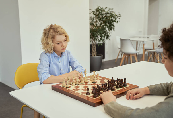 Harekete geçmeden önce düşün. Düşünceli küçük beyaz çocuk arkadaşıyla satranç oynuyor, okulda masada birlikte oturuyor. - Fotoğraf, Görsel