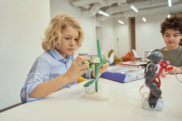 Dinge erschaffen. Kaukasischer Junge untersucht technisches Spielzeug, während er mit anderen Kindern am Tisch sitzt und Ingenieurunterricht hat - Foto, Bild