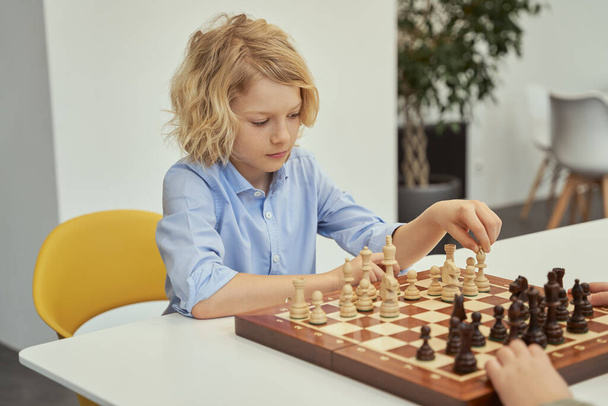 Busco un buen movimiento. Curioso niño caucásico en camisa azul sentado en el aula y jugando ajedrez en el tablero de ajedrez - Foto, imagen