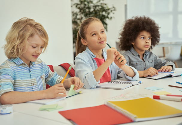 Az oktatás kulcsfontosságú. Kedves, változatos gyerekek tanulnak és jegyzetelnek, miközben együtt ülnek az asztalnál az általános iskolai osztályteremben. - Fotó, kép