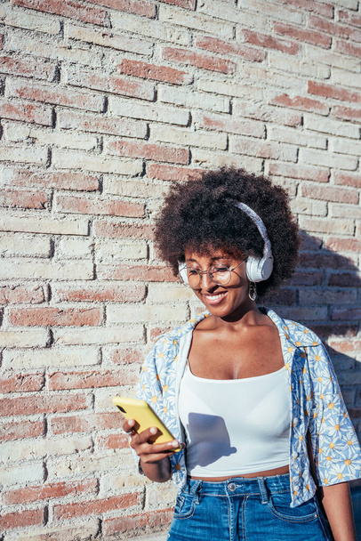 retrato vertical de uma jovem africana com seu telefone e fones de ouvido na rua - Foto, Imagem