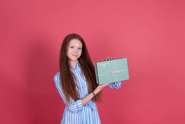 Mavi elbiseli 13 yaşındaki küçük kız pembe arka planda izole edilmiş, elinde hediye kutusu mutlu bir şekilde gülümsüyor. - Fotoğraf, Görsel