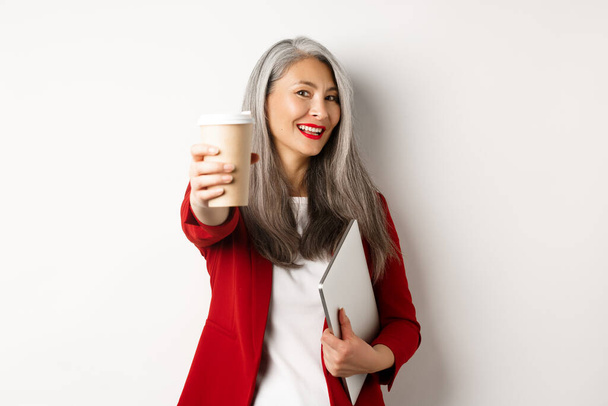 По делам. Веселая азиатская женщина менеджер давая вам чашку кофе и улыбаясь, стоя с ноутбуком в руке, белый фон - Фото, изображение