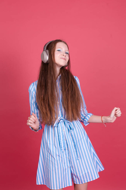 Little kid girl 13 years old in blue dress with brackets isolated on pink background in wireless headphones happy smiling dancing moving - Photo, Image