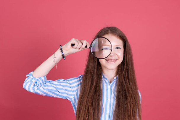 Little kid girl 13 years old in blue dress isolated on pink background does grimaced in magnifier - Photo, Image