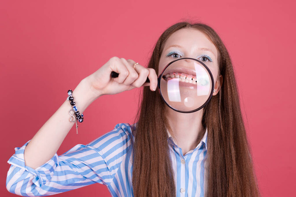 Niña de 13 años en vestido azul aislada sobre fondo rosa muestra sus dientes con paréntesis en lupa - Foto, Imagen