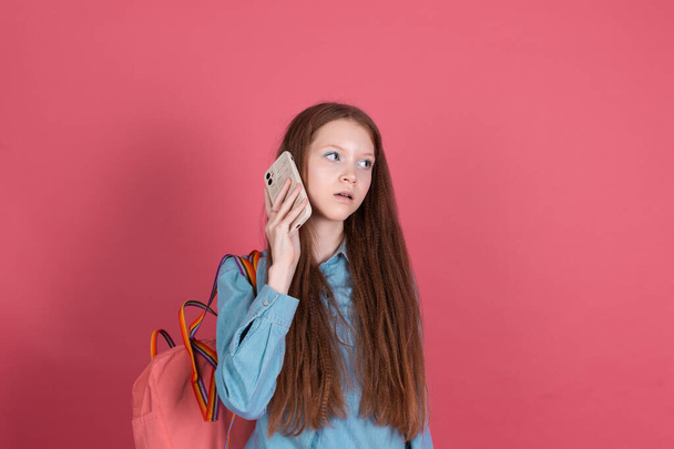 Niña 13 años aislada en fondo rosa colegiala con mochila con teléfono móvil aburrido escuchar mensaje de voz, tener conversación - Foto, imagen