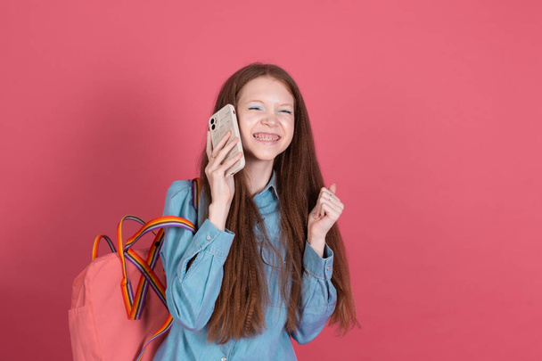 Κοριτσάκι 13 ετών σε μπλε τζιν μπουφάν απομονωμένο σε κόκκινο φόντο μαθήτρια με σακίδιο πλάτης με κινητό τηλέφωνο χαρούμενη χειρονομία νικητής - Φωτογραφία, εικόνα