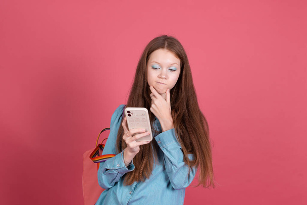 Маленькая девочка 13 лет в синей джинсовой куртке изолирована на красном фоне школьница с рюкзаком с мобильным телефоном вдумчивый взгляд на экране - Фото, изображение