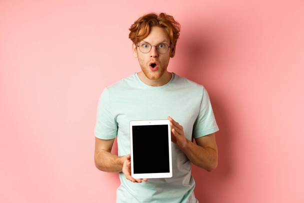 Excited young man with red hair and beard, checking out online promotion, showing digital tablet screen and staring at camera amazed, standing over pink background - Photo, Image