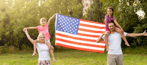 Eltern und Kind mit amerikanischer Flagge spielen mit einem bunten Drachen. Mutter, Vater und ihre kleinen Töchter feiern bei nebligem Wetter gemeinsam im Freien. Konzept zum Unabhängigkeitstag der USA. - Foto, Bild