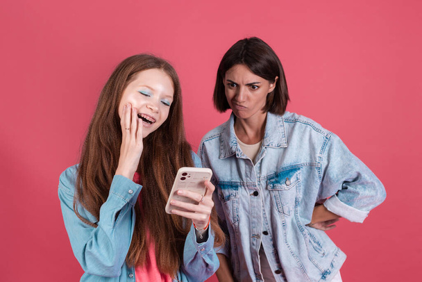 Modern mom and daughter in denim jackets on terracotta background, girl  with phone and mom stands by with reproachful gaze - Photo, image