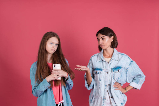 Modern mom and daughter in denim jackets on terracotta background, girl  with phone and mom stands by trying to take phone - Photo, Image
