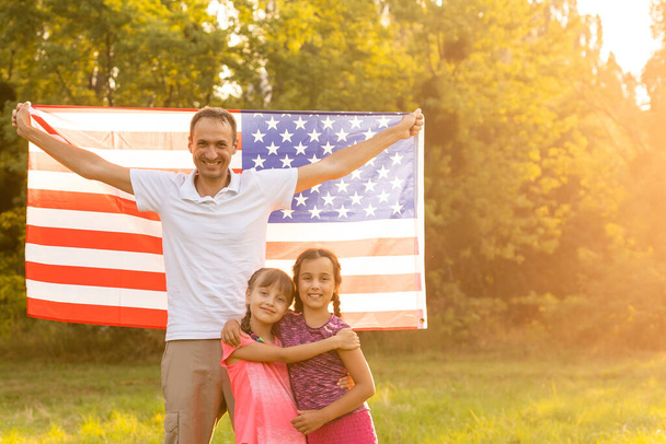 Happy family sitting together in their backyard holding the american flag behind them. Smiling couple with their kids celebrating american independence day holding american flag - Photo, Image