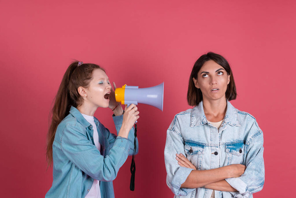 Modern mom and daughter in denim jackets on terracotta background girl shouting in megaphone mom stand by with bored dissatisfied gaze - Foto, Imagem