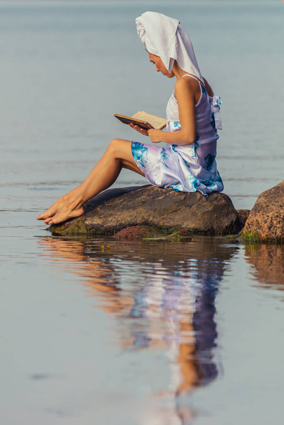 Молодая женщина с полотенцем на голове у моря. Леди читает книгу в воде. Концепция летнего отдыха на открытом воздухе, релаксации. чтение - Фото, изображение