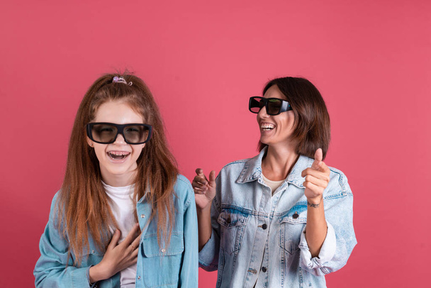 Modern mom and daughter in denim jackets on terracotta background in 3 d cinema glasses watching comedy movie together, smile and laugh - Photo, Image