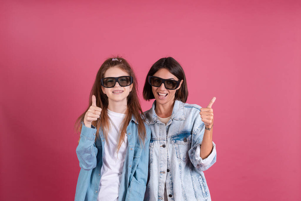 Modern mom and daughter in denim jackets on terracotta background in 3d cinema glasses positive smiling shows thumb up - Photo, Image