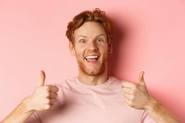 Крупный план веселого человека с рыжими волосами и бородой, показывая большие пальцы вверх и улыбаясь, говоря да, одобрить и похвалить что-то прохладное, стоя на розовом фоне - Фото, изображение