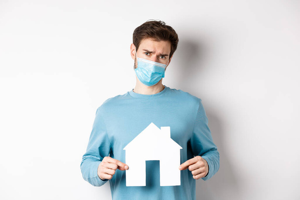 Covid and real estate concept. Sad and doubtful young man in medical mask feeling reluctant, showing paper home cutout, standing over white background - Photo, Image