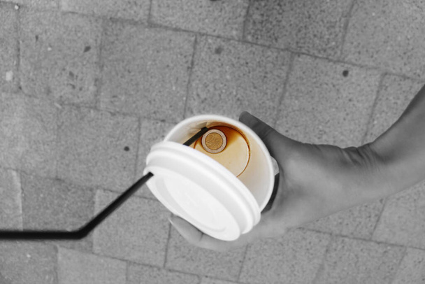 A two euro coin at the bottom of a paper coffee cup. Leftover coffee and a plastic tube. Hold a glass in your hand against a background of paving stones. - Photo, Image
