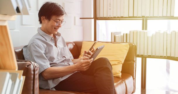 Senior Aziatische man speelt tablet en video oproep op de bank in de woonkamer thuis Portret van Aziatische oudere man is ontspannen en gelukkig Met het spelen van tablet - Foto, afbeelding