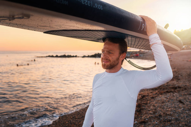 Portret van een blanke man die een bord tegen zijn hoofd houdt en over het strand loopt. Op de achtergrond, de oceaan en de zonsondergang. Zomer extreme recreatie en surfen. - Foto, afbeelding