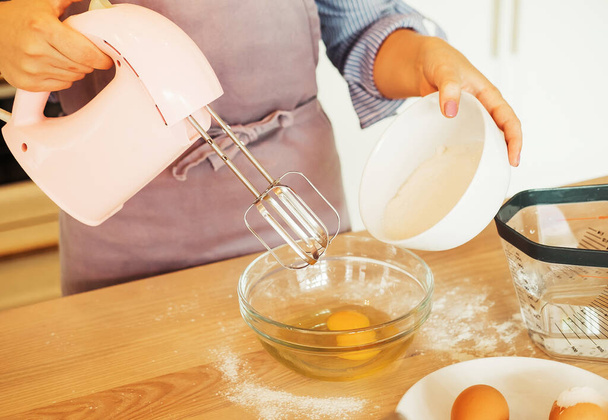 Young woman baking a cake in the kitchen, using a mixer to whisk the fresh ingredients in a glass mixing bowl. - Foto, immagini