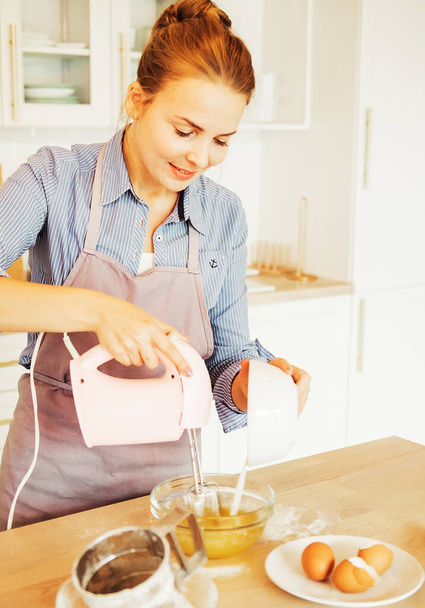 Giovane donna che cuoce una torta in cucina, utilizzando un mixer per sbattere gli ingredienti freschi in una ciotola di vetro. - Foto, immagini