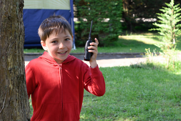 Porträt eines Kindes, das in die Kamera blickt. Der Junge hält einen Walkie Talkie in der Hand und befindet sich auf einem Campingplatz - Foto, Bild