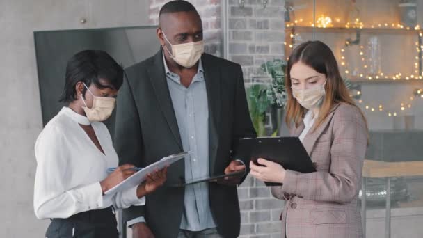 Multiethnic business team colleagues partners two multiracial women and african mature boss man leader wear face masks stand in company office discuss project read papers documents report strategy - Footage, Video