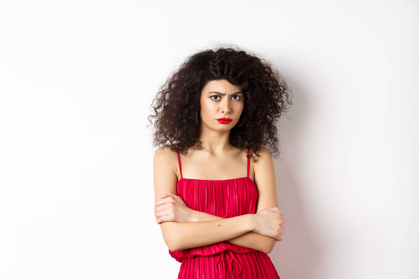 Sad and offended caucasian woman in red dress frowning, cross arms on chest and sulking, feeling mad at you, standing over white background - Foto, afbeelding
