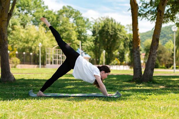 Yoga. Eine junge Frau steht in der Pose eines Hundes, der nach unten schaut, und macht Yoga im Park auf dem Rasen. Das Konzept von gesundem Lebensstil und Sport. - Foto, Bild